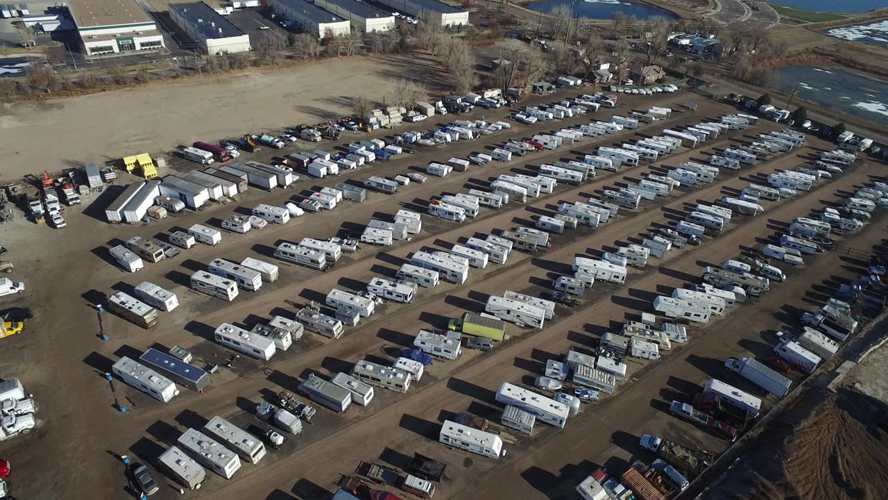 Opening aerial frame over a Hidden Lake, Colorado dirt vehicle lot with rows of RVs and cars, low industrial buildings beyond the lot, and no visible people.
