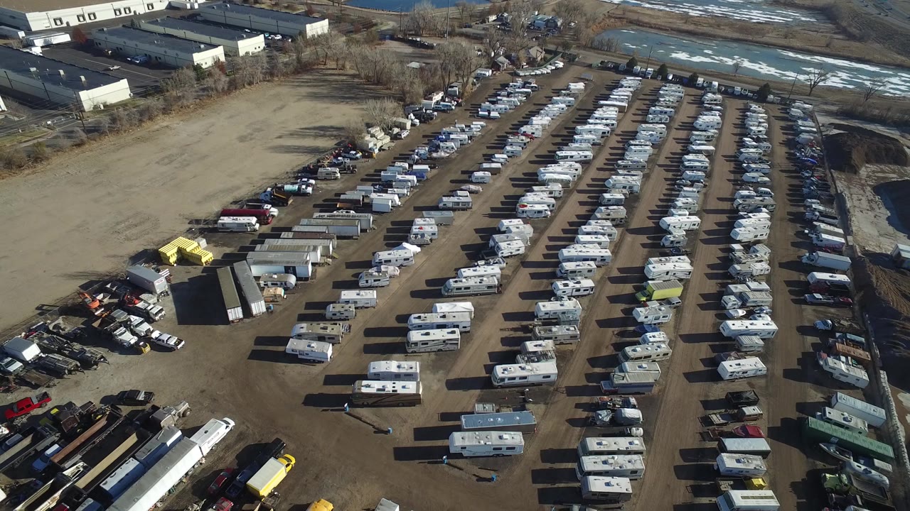 Aerial frame in Hidden Lake, Colorado with white RVs and trailers lined across a dirt yard, industrial buildings at the top edge, and no visible people.