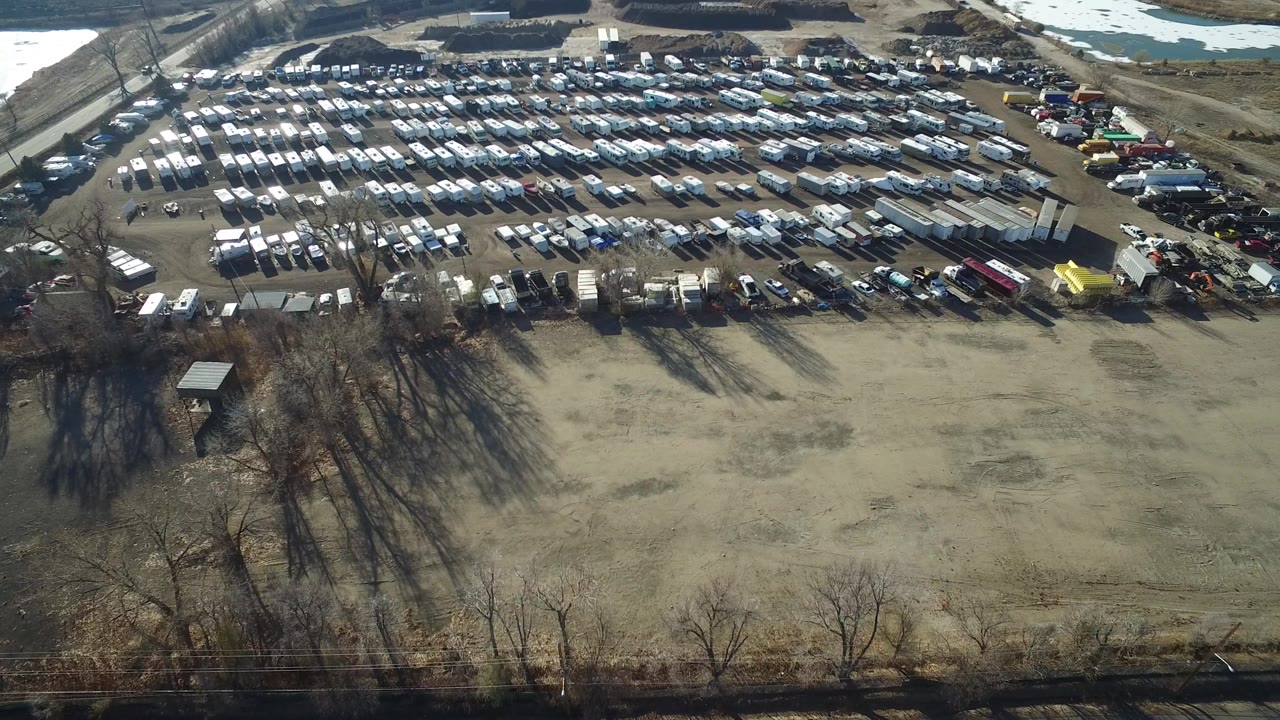 Aerial view in Hidden Lake, Colorado showing dense rows of parked cars and light vehicles beyond a bare open lot, with leafless trees and patches of snow near the top edge.