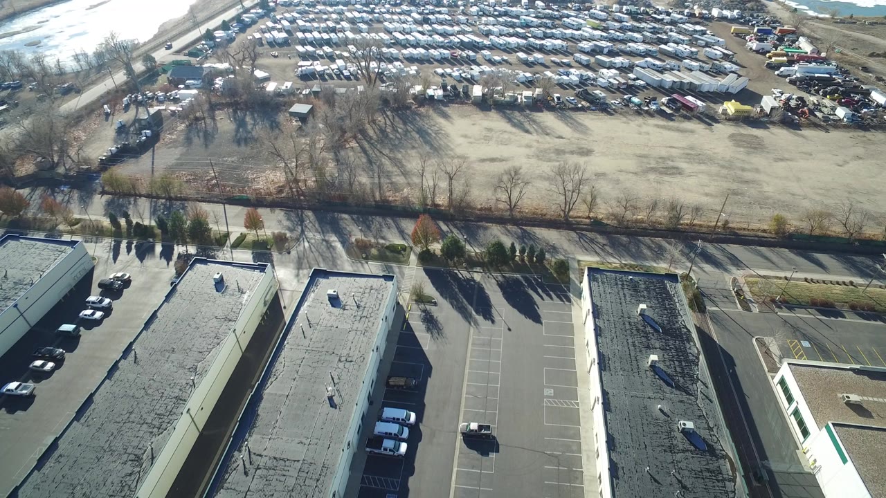 Aerial frame in Hidden Lake, Colorado showing two long warehouse-style buildings, paved lots with a few vehicles, bare trees, and the vehicle yard beyond, with no visible people.