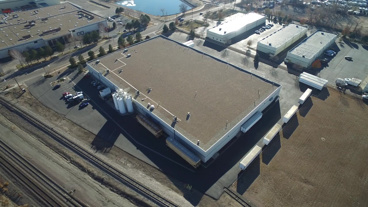 Aerial frame from Hidden Lake, Colorado showing a flat-roof warehouse beside several rail tracks, with loading bays, white storage silos, parked cars, and no visible people.