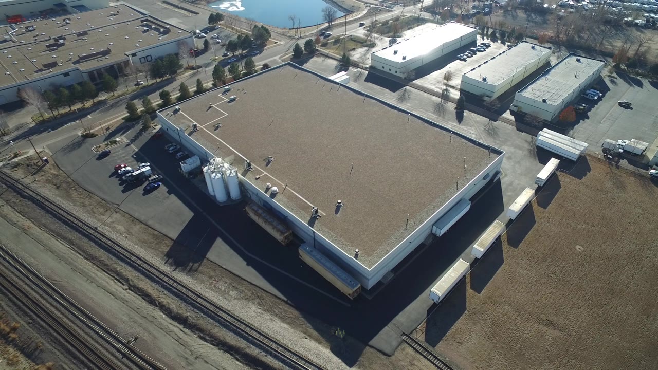 Aerial view in Hidden Lake, Colorado with the warehouse centered, multiple rail lines along the lower edge, small industrial buildings beyond the road, and no visible people.