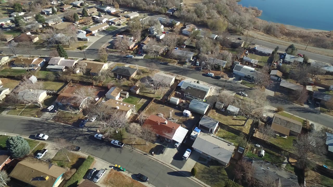 Aerial frame over Hidden Lake, Colorado with homes, driveways, street corners, bare trees, and the lake visible beyond the neighborhood, no visible people.