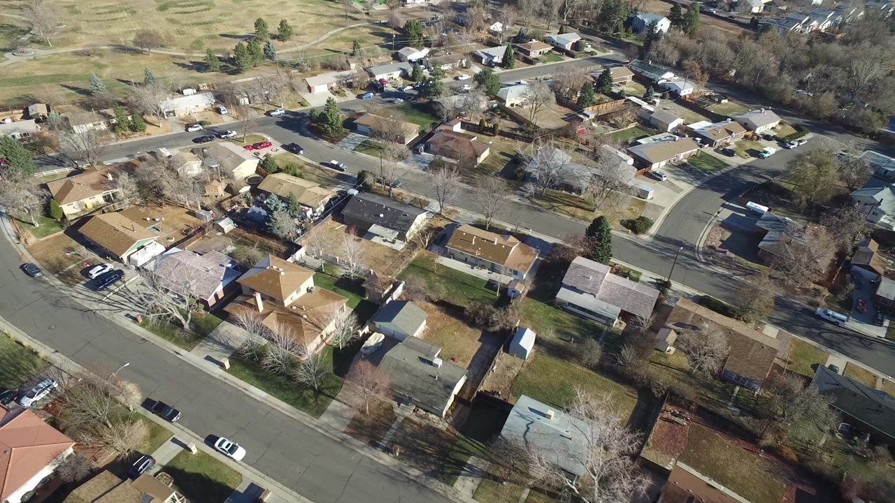 High aerial frame over Hidden Lake, Colorado showing detached homes, curved neighborhood streets, leafless trees, parked vehicles, and open grassy fields in the background, with no visible people.