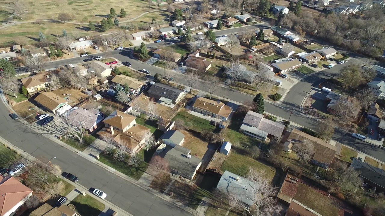 Oblique aerial view in Hidden Lake, Colorado with tan and brown rooftops, residential roads, small yards, bare trees, parked cars, and open land beyond the houses, with no visible people.