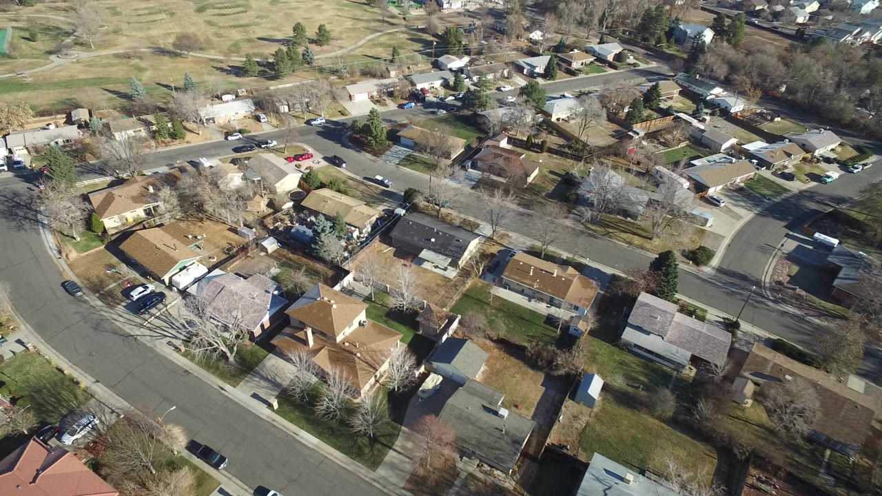 Drone-like aerial over suburban homes, roads, trees, and a park field.