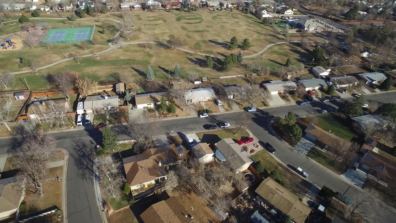 Elevated drone view in Hidden Lake, Colorado showing rooftops and yards below, a residential road through the middle, and park paths leading toward tennis courts with no recognizable people.
