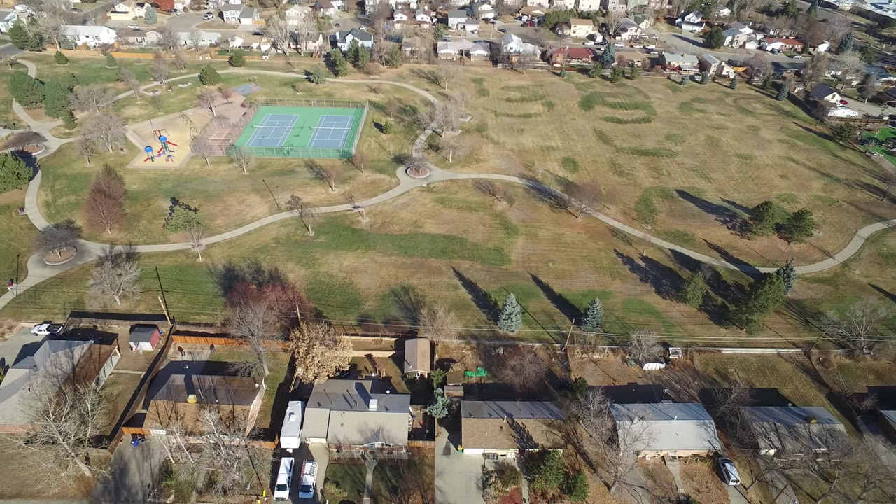 High aerial frame over Hidden Lake, Colorado homes, a street, winter-brown park grass, curved paths, playground equipment, and green tennis courts, with no visible people.