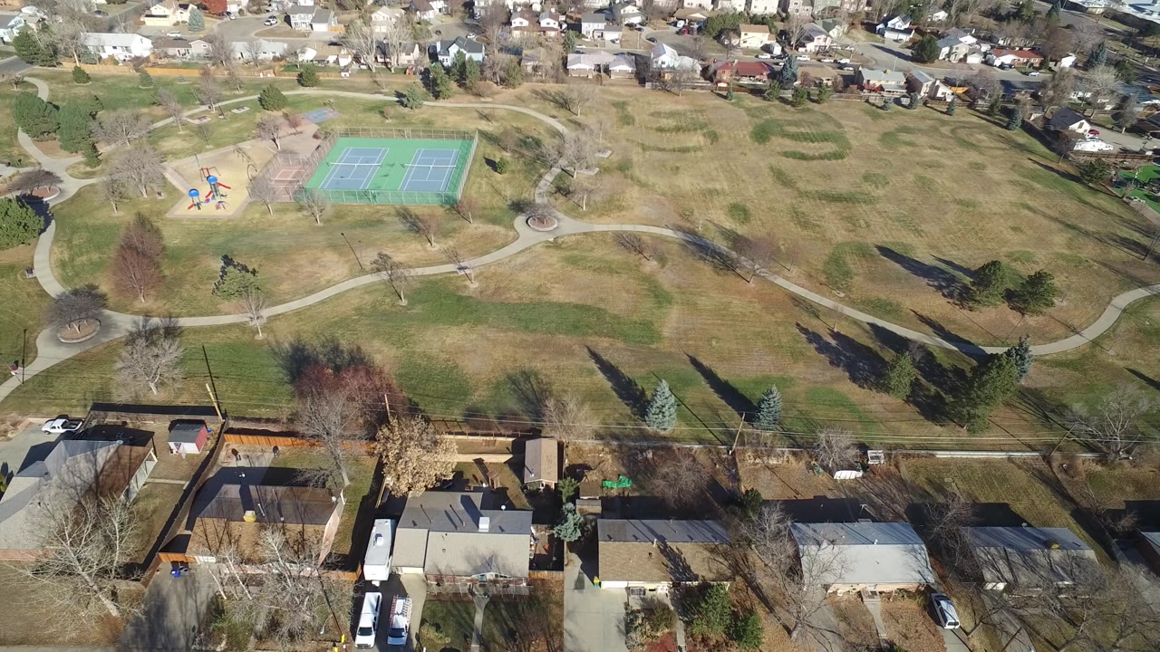 Aerial view in Hidden Lake, Colorado with foreground rooftops and parked vehicles below a neighborhood park, playground, walking paths, and tennis courts, with no visible people.
