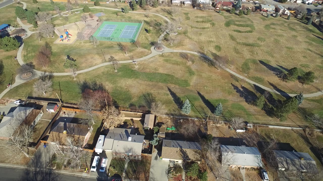 Aerial view in Hidden Lake, Colorado, with houses and parked vehicles along the lower edge, a road through the neighborhood, park lawns, bare trees, a playground, and green tennis courts beyond.