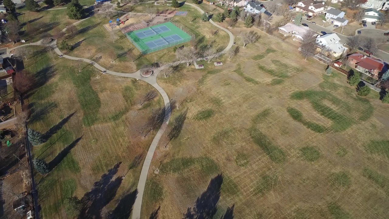 Aerial view of a park with curved paths, tennis courts, trees, and nearby homes.