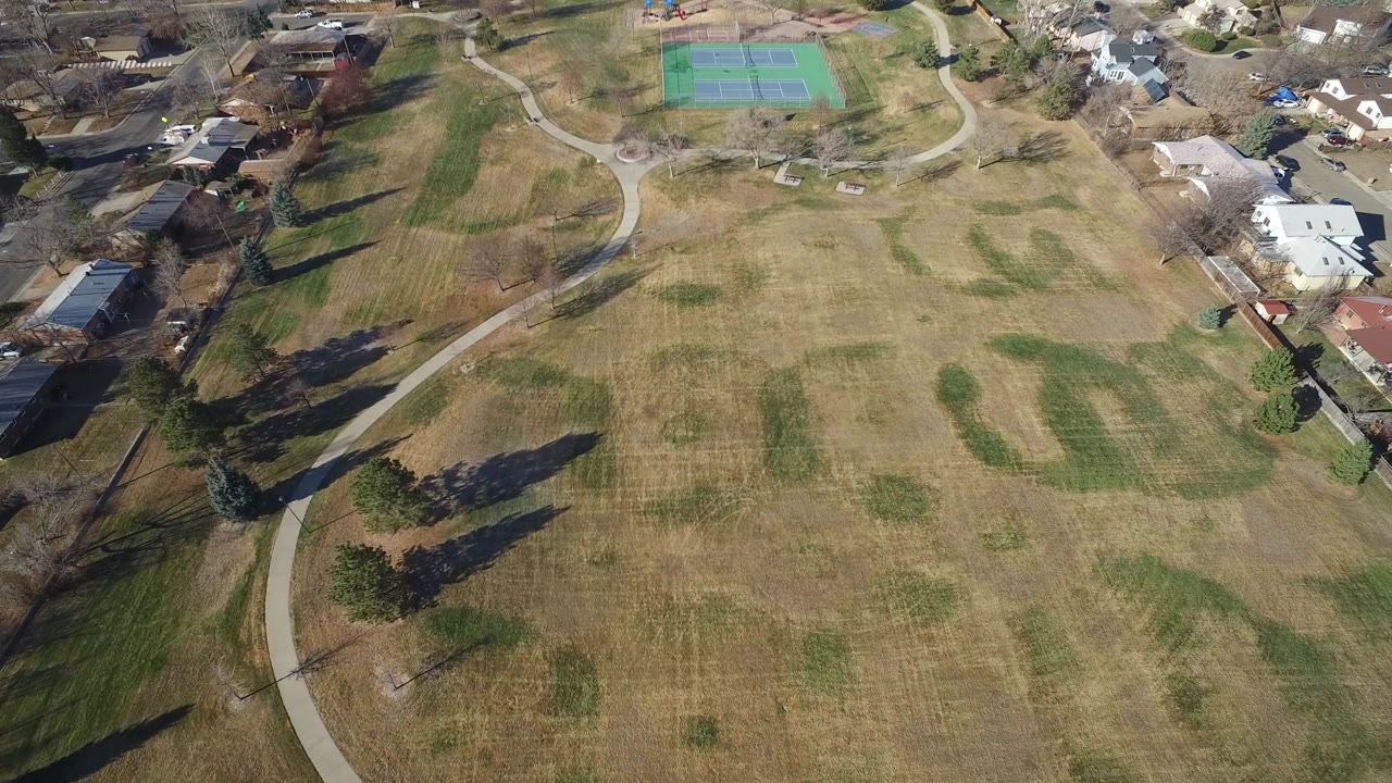 Aerial frame over Hidden Lake, Colorado showing a large brown-green field, curving paths, nearby homes, tennis courts at the top, and no visible people.