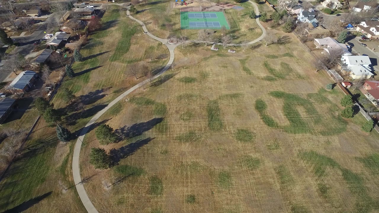 High aerial view in Hidden Lake, Colorado with the field centered, a curving path along the left, residential buildings along the top and right, tennis courts near the top edge, and no visible people.