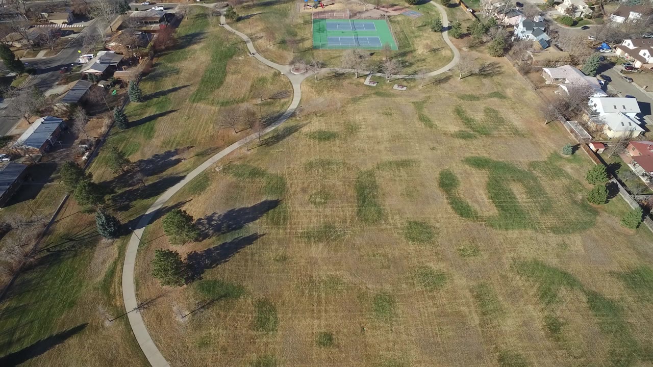 Drone view of patchy grass, paths, homes, and green tennis courts.