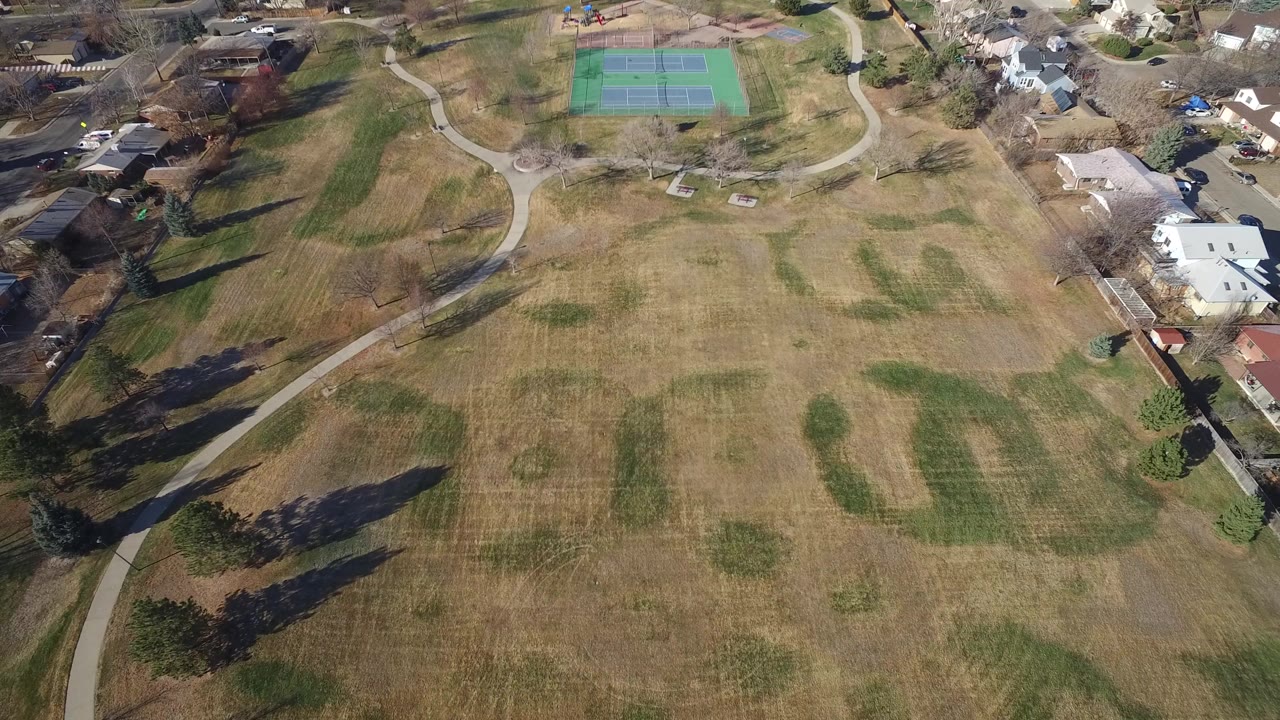 Aerial view of a broad park field with paths, tennis courts, trees, and nearby houses.
