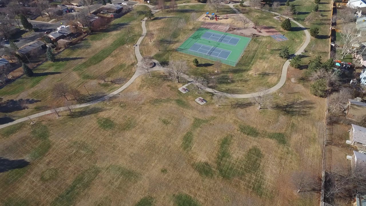 Aerial view in Hidden Lake, Colorado with the tennis courts slightly right of center, broad winter-colored grass in the foreground, a circular path feature, homes, trees, and no visible people.