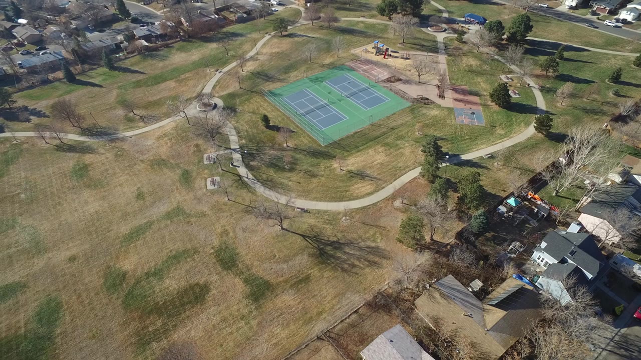 High aerial frame over a Hidden Lake, Colorado park with green tennis courts near the top center, curving paths through open grass, houses at the lower right, and no visible people.