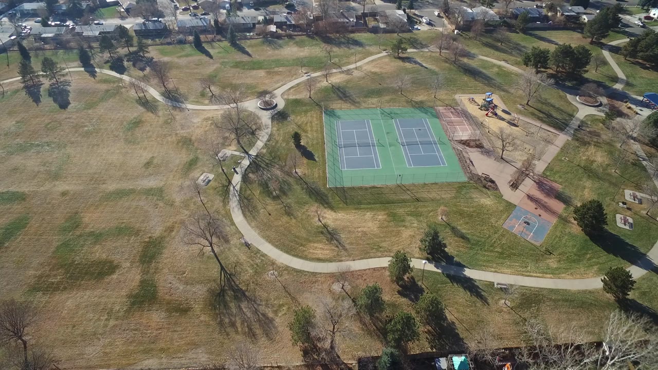 Aerial view in Hidden Lake, Colorado with two tennis courts centered lower in frame, a curving path crossing the grass, scattered trees, and residential roofs beyond the park.