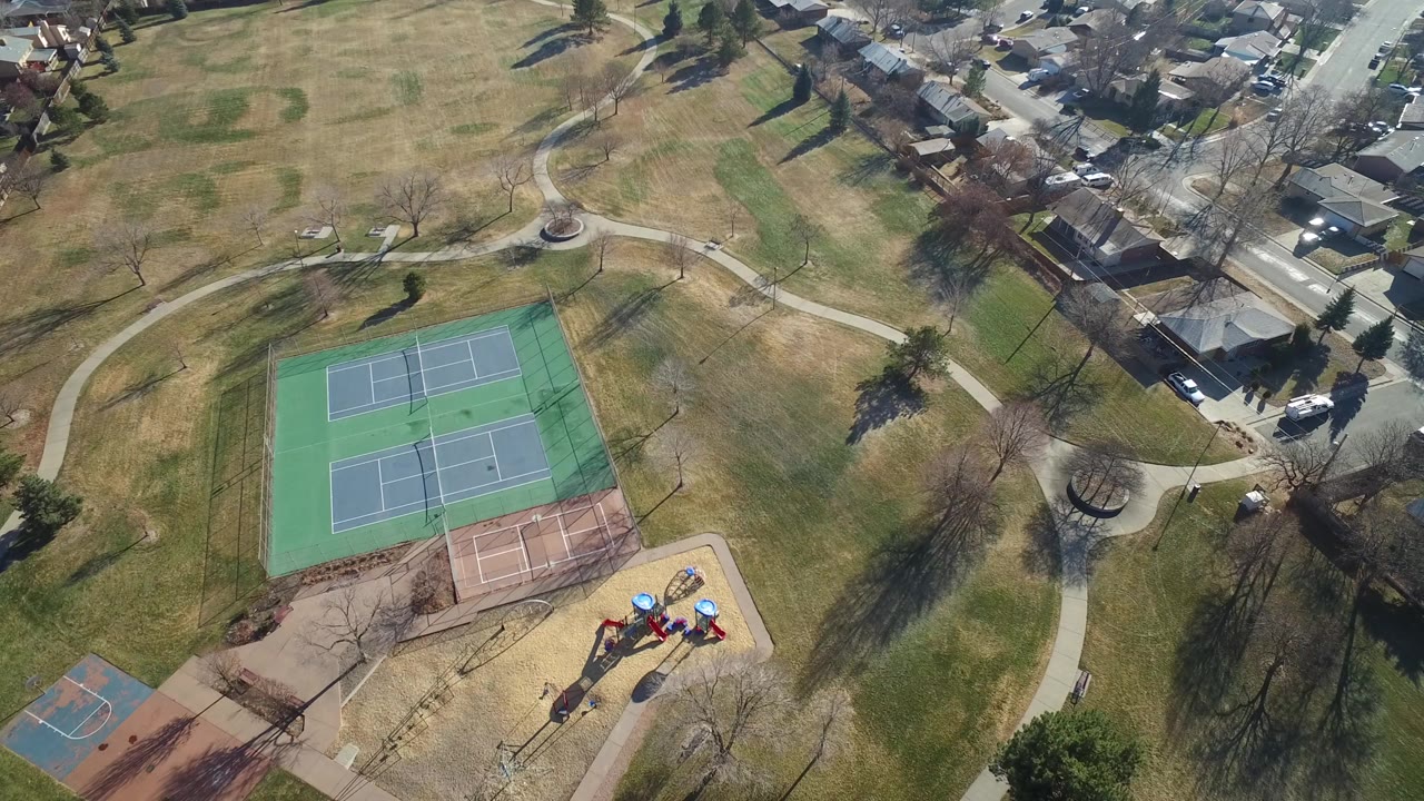 Aerial frame over Hidden Lake, Colorado parkland showing tennis courts, a playground, walking paths, bare trees, open grass, and nearby residential streets.