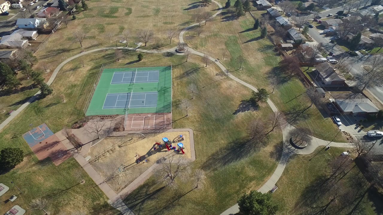 High aerial frame over Hidden Lake, Colorado showing green tennis courts, a reddish court, a playground, curving paths, open grass, nearby houses, and no visible people.
