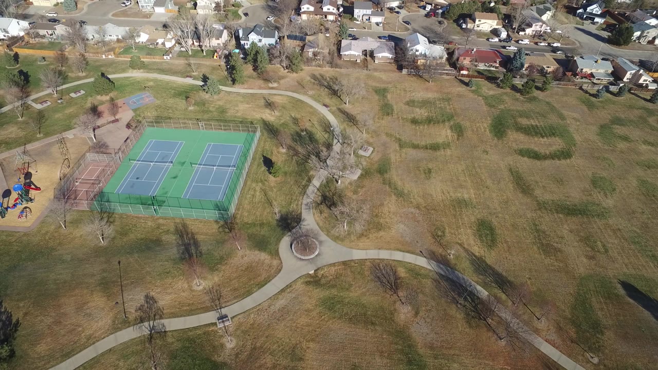 Aerial view of fenced tennis courts, park paths, lawns, playground, and nearby homes.