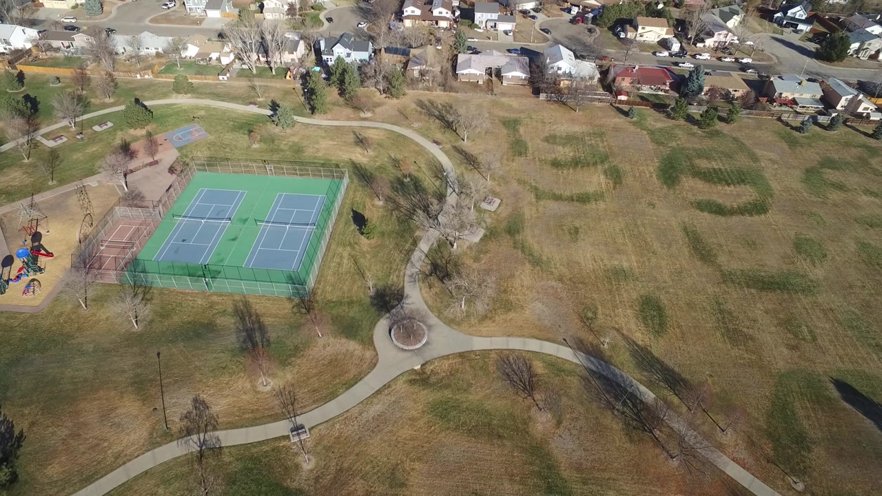 Aerial view in Hidden Lake, Colorado with the tennis courts left of center, open brown-green park grass across the middle, winding paths, houses at the top, and no visible people.