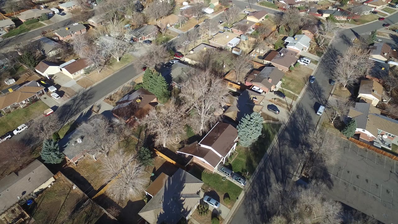 Aerial frame over Hidden Lake, Colorado showing rooftops, fenced backyards, a broad street junction, winter-bare trees, and several parked vehicles, with no clearly identifiable people visible.