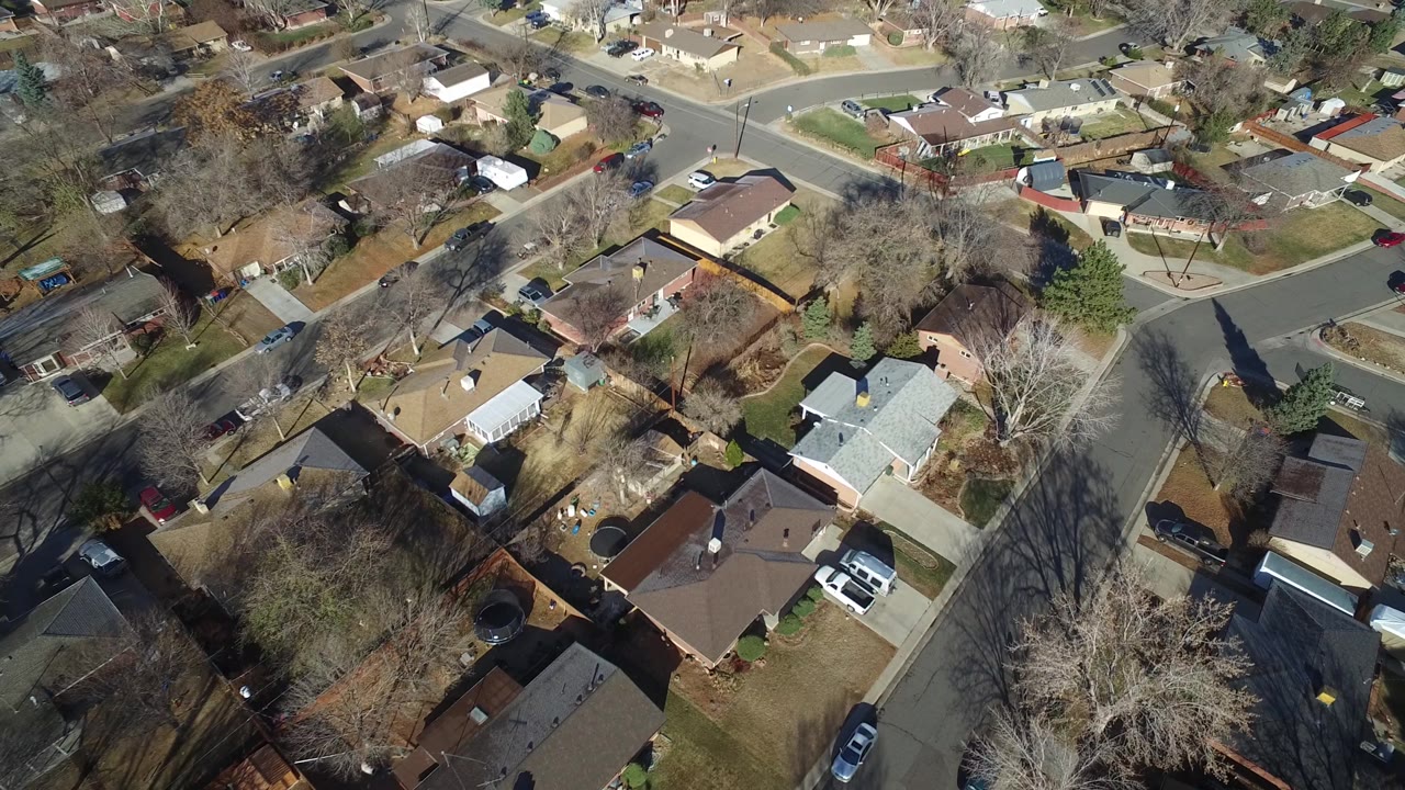 Aerial frame over Hidden Lake, Colorado rooftops, fenced backyards, a residential intersection, parked vehicles, and winter-bare trees under bright sun.