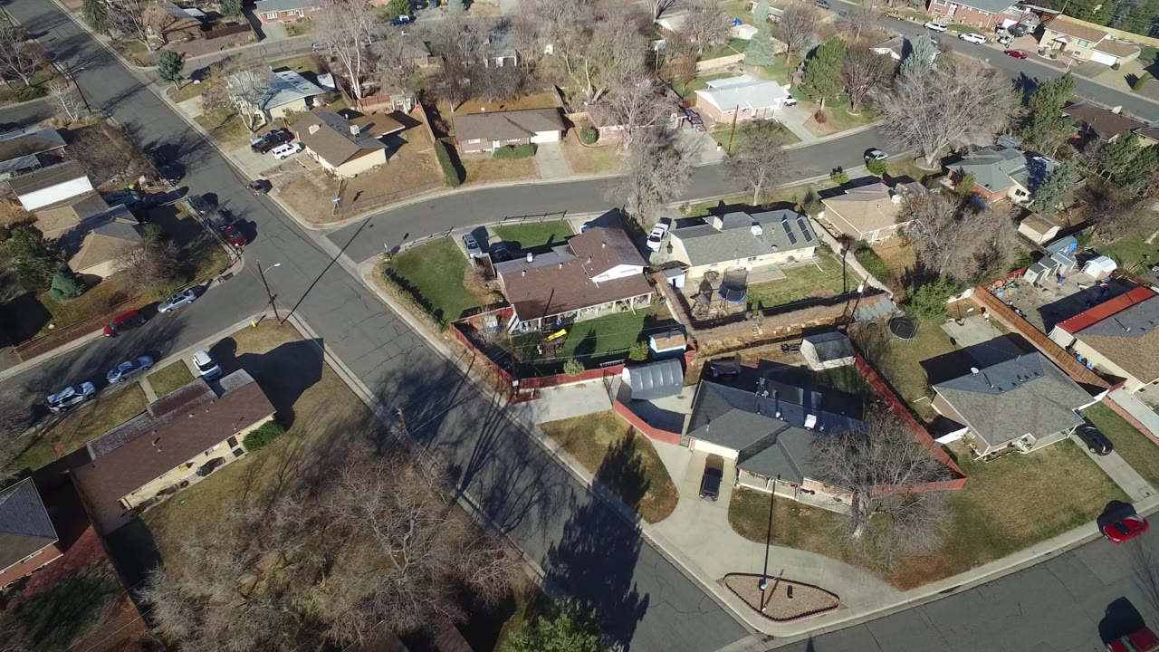 Drone-height frame over Hidden Lake, Colorado with a curved street, rooftops, fenced backyards, lawns, parked vehicles, bare trees, and no visible people.