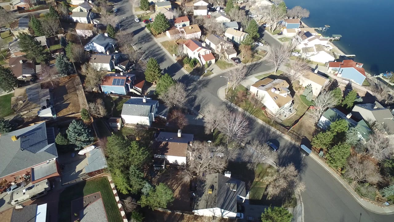 Aerial view over Hidden Lake, Colorado homes with curved residential streets, leafless trees, parked vehicles, and the lake edge at the upper right, with no visible people.