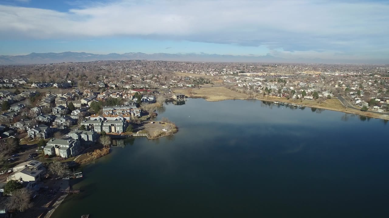 Aerial frame in Hidden Lake, Colorado showing the left shoreline buildings more centrally, broad water at bottom, a spread of houses beyond the lake, hazy hills, cloud bands, and no visible people.
