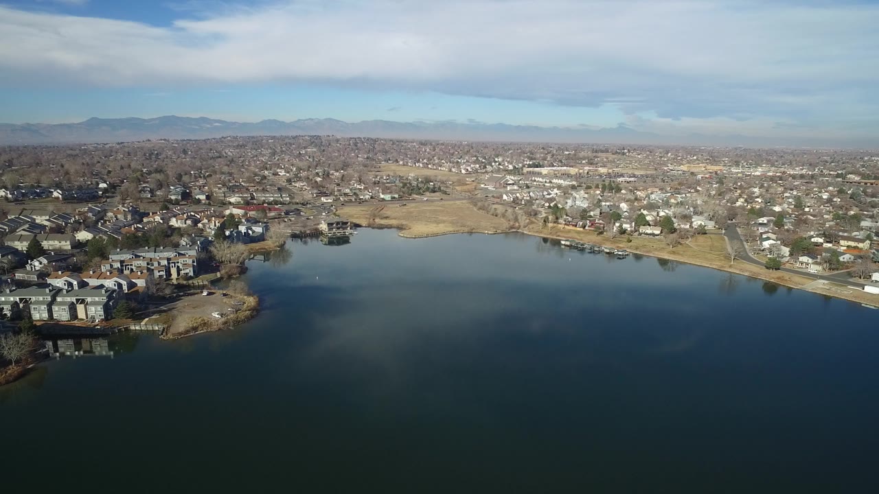 Aerial view over dark blue lake water toward homes and distant mountains.