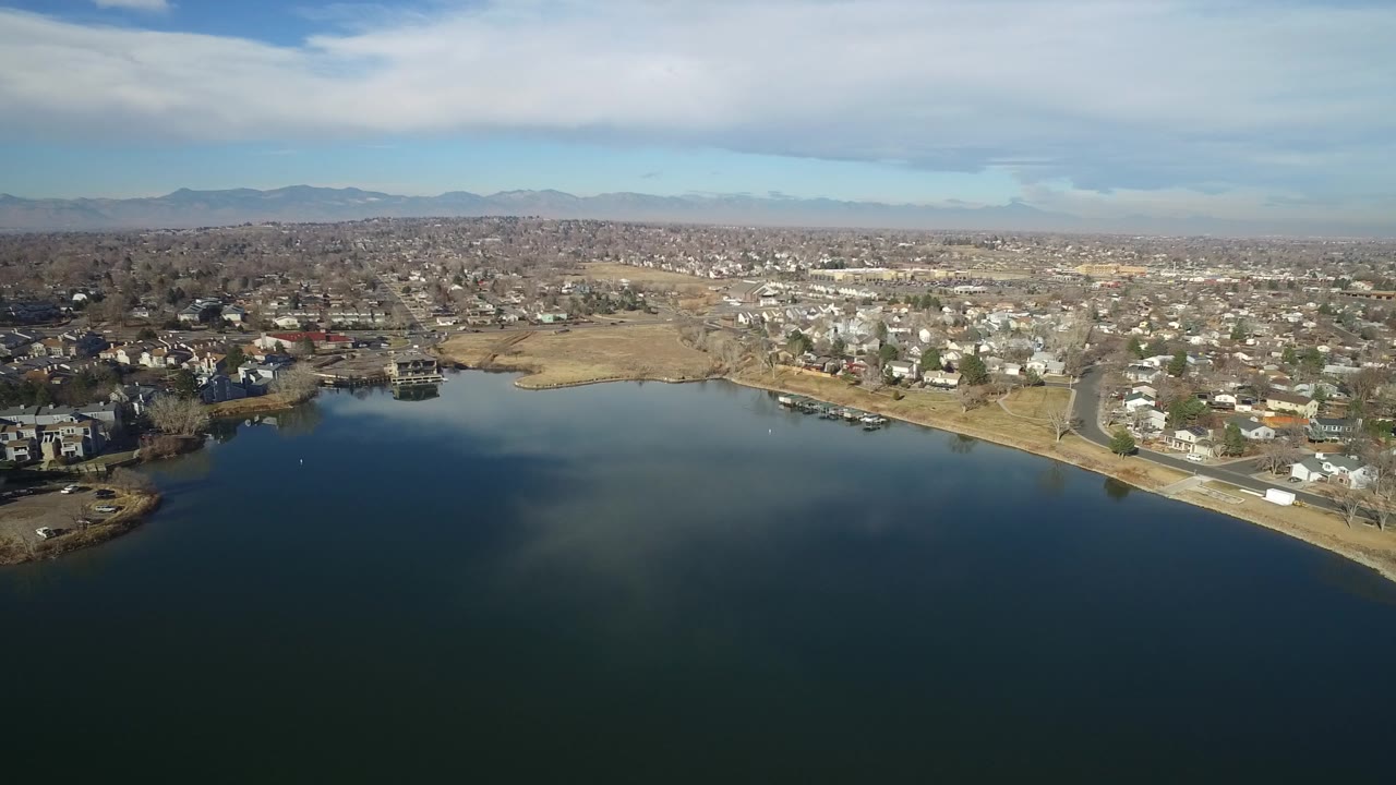 Aerial view over dark lake water toward shoreline homes and distant mountains.