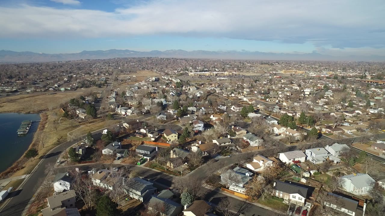 Aerial view of suburban homes, yards, roads, lake edge, and distant mountains.