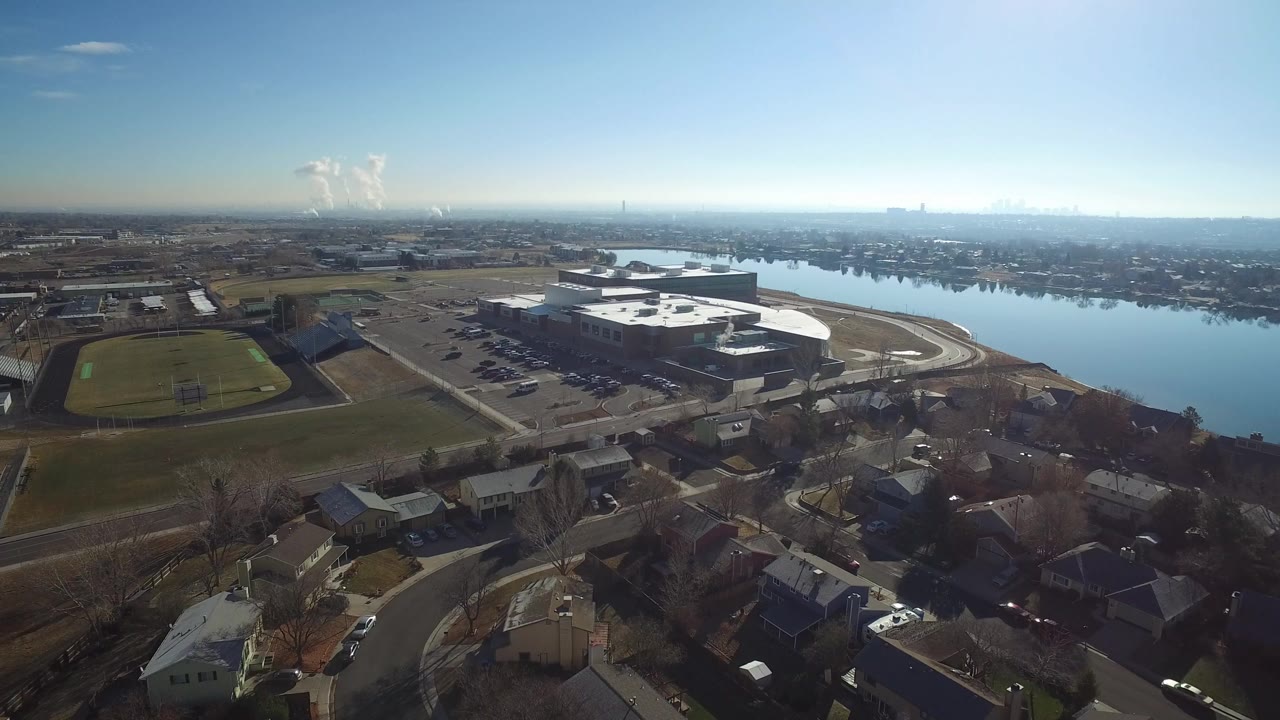 Aerial view of homes, a sports field, a large lakeside building, and a lake under clear sky.