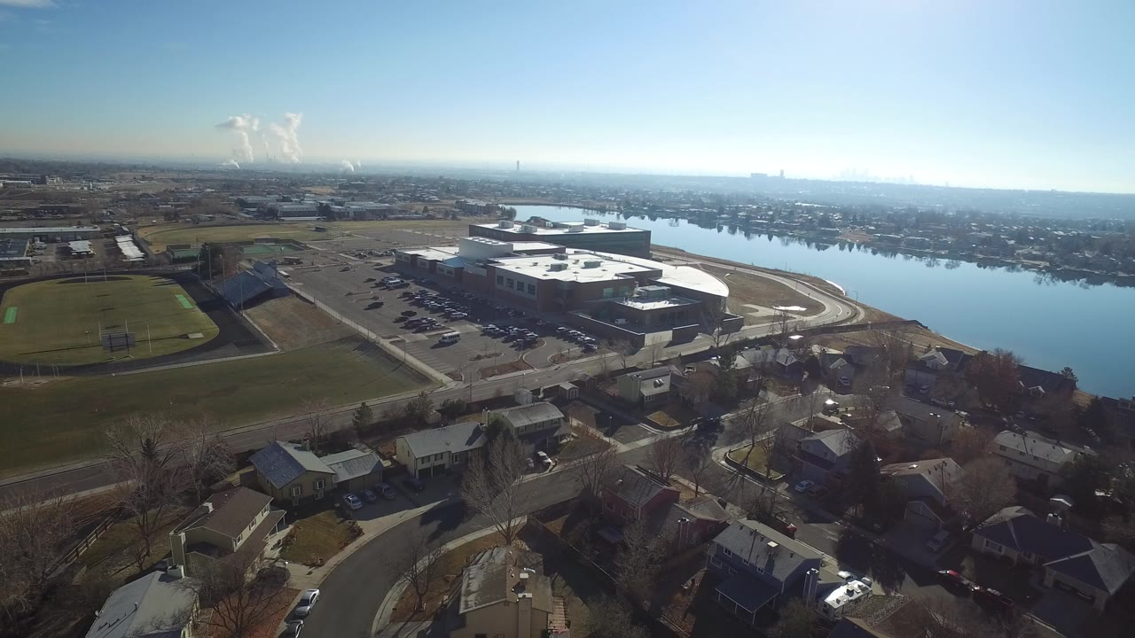 Aerial view of a sports field, large building, parking lot, homes, and waterway.
