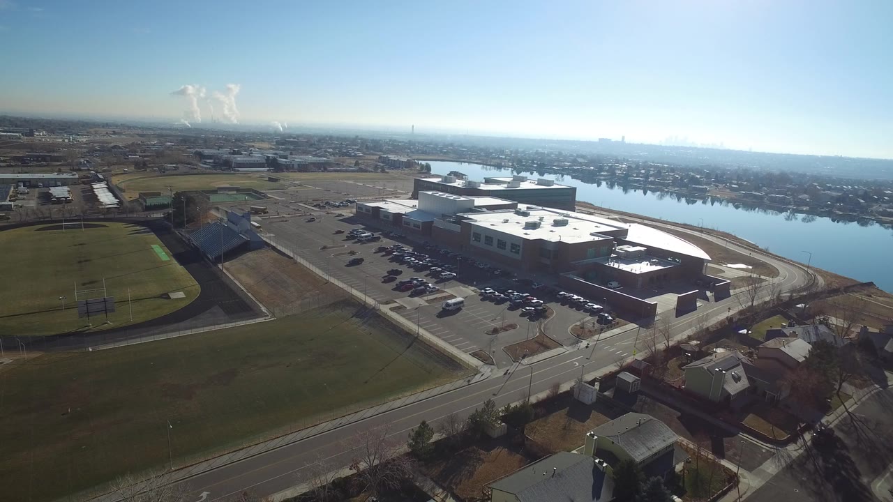 Elevated aerial view in Hidden Lake, Colorado, with a corporate building and parking lot beside a soccer field, reflective water in the background, and no visible people.