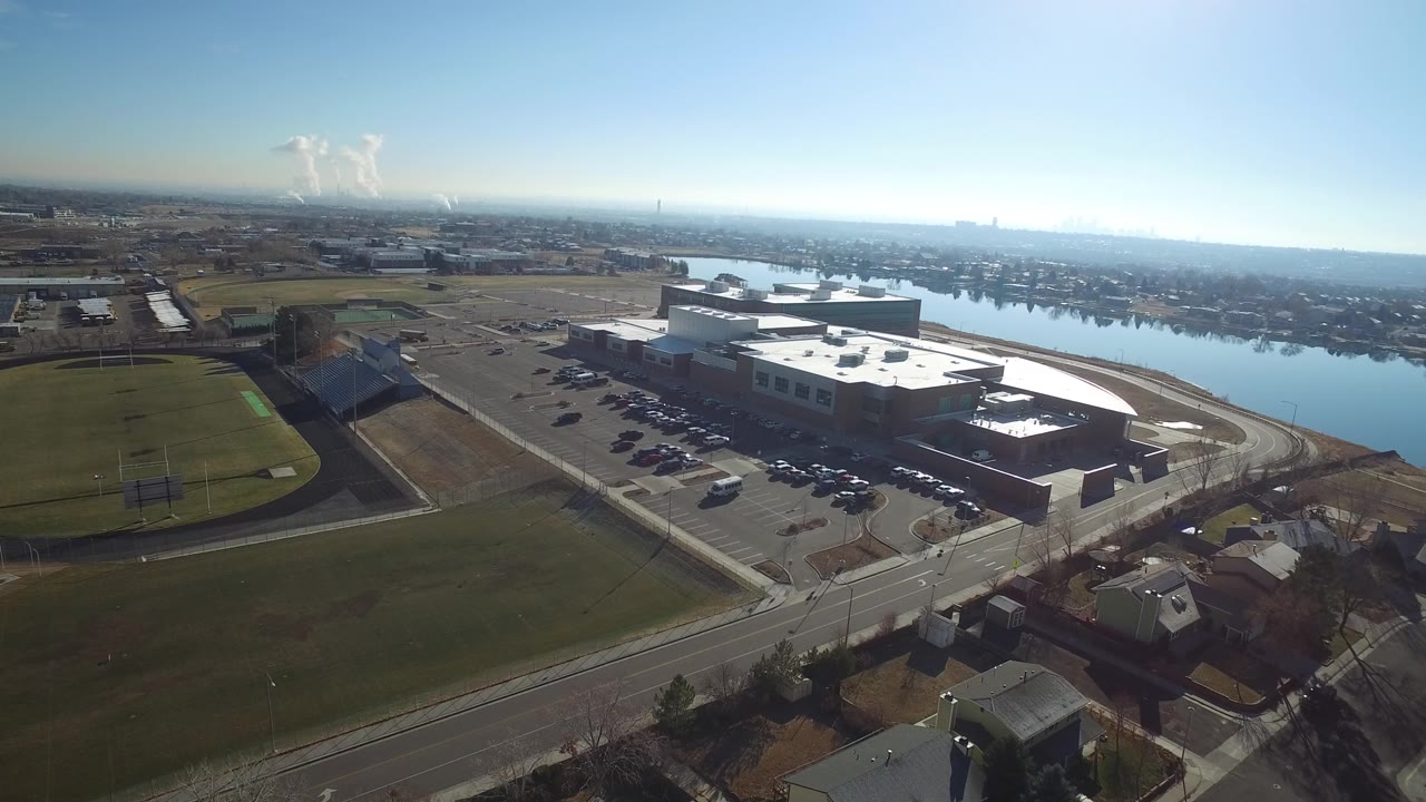 Hidden Lake, Colorado aerial frame showing the low-rise building slightly closer, with rows of parked cars, green playing fields, water, horizon haze, and no visible people.