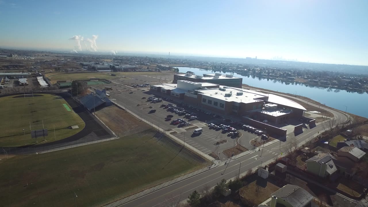 Drone-like aerial of a large building beside water with sports fields in the foreground.