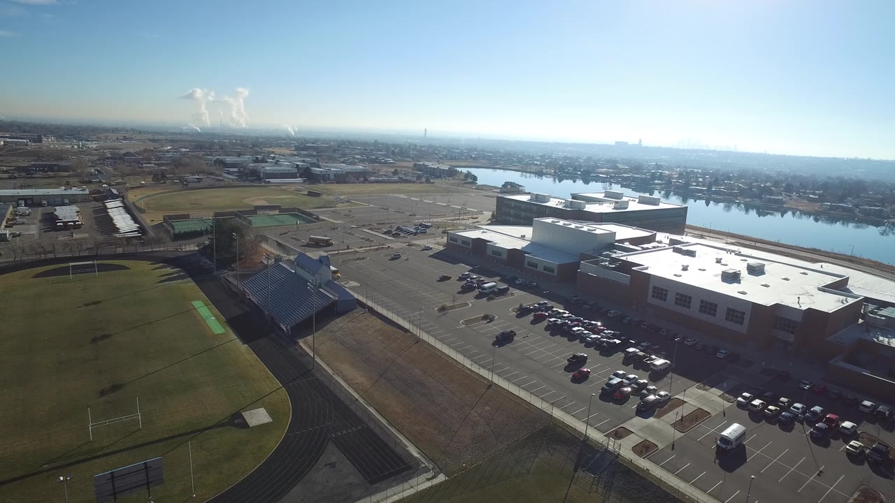Aerial view in Hidden Lake, Colorado with bleachers lower left, green playing fields, a large lot of parked cars, flat-roofed buildings, river water, and no visible people.