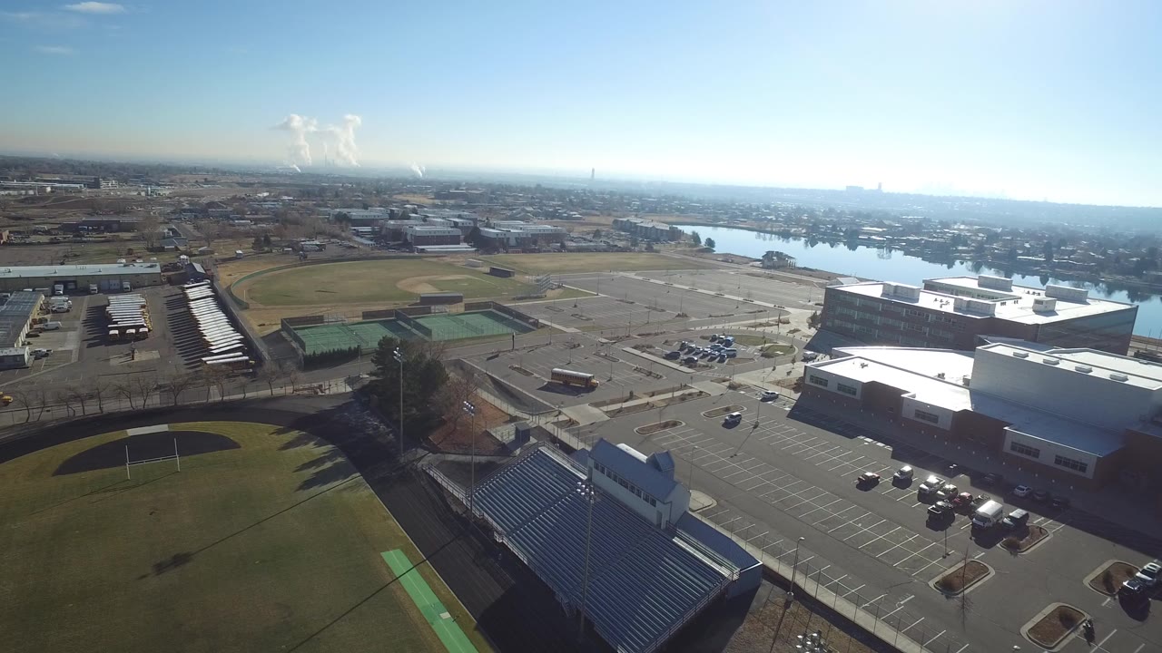 Aerial frame in Hidden Lake, Colorado showing baseball diamonds, green tennis courts, large paved parking areas, low industrial buildings, distant river water, and no visible people.