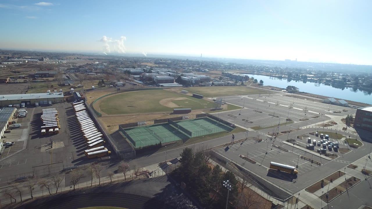 High aerial frame over Hidden Lake, Colorado, showing tennis courts, a baseball field, rows of buses, a large parking lot, distant water, and no visible people.