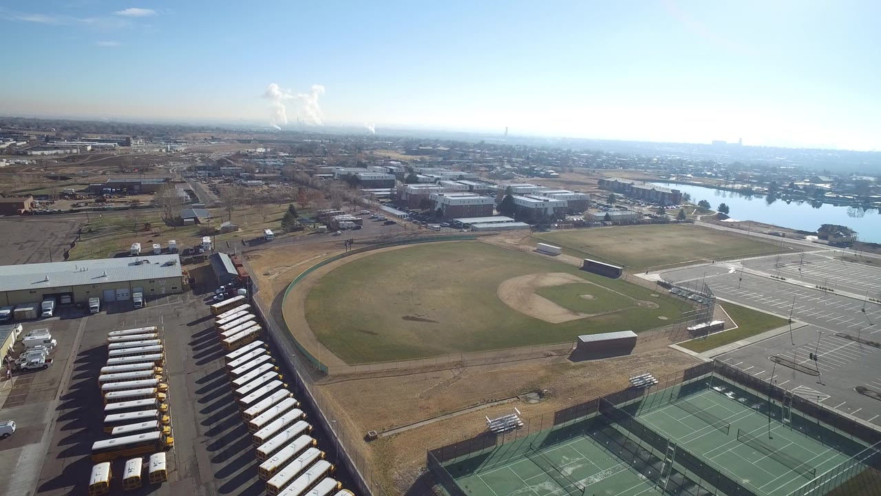 Aerial view over tennis courts, a baseball field, parking areas, low buildings, and a lake in the distance.