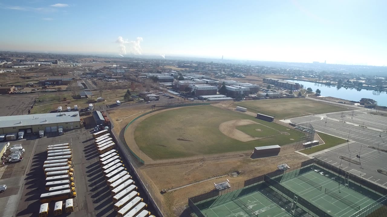 Early aerial frame in Hidden Lake, Colorado with the baseball diamond centered, tennis courts lower right, parked buses or trailers at lower left, lake water behind the buildings, and no visible people.