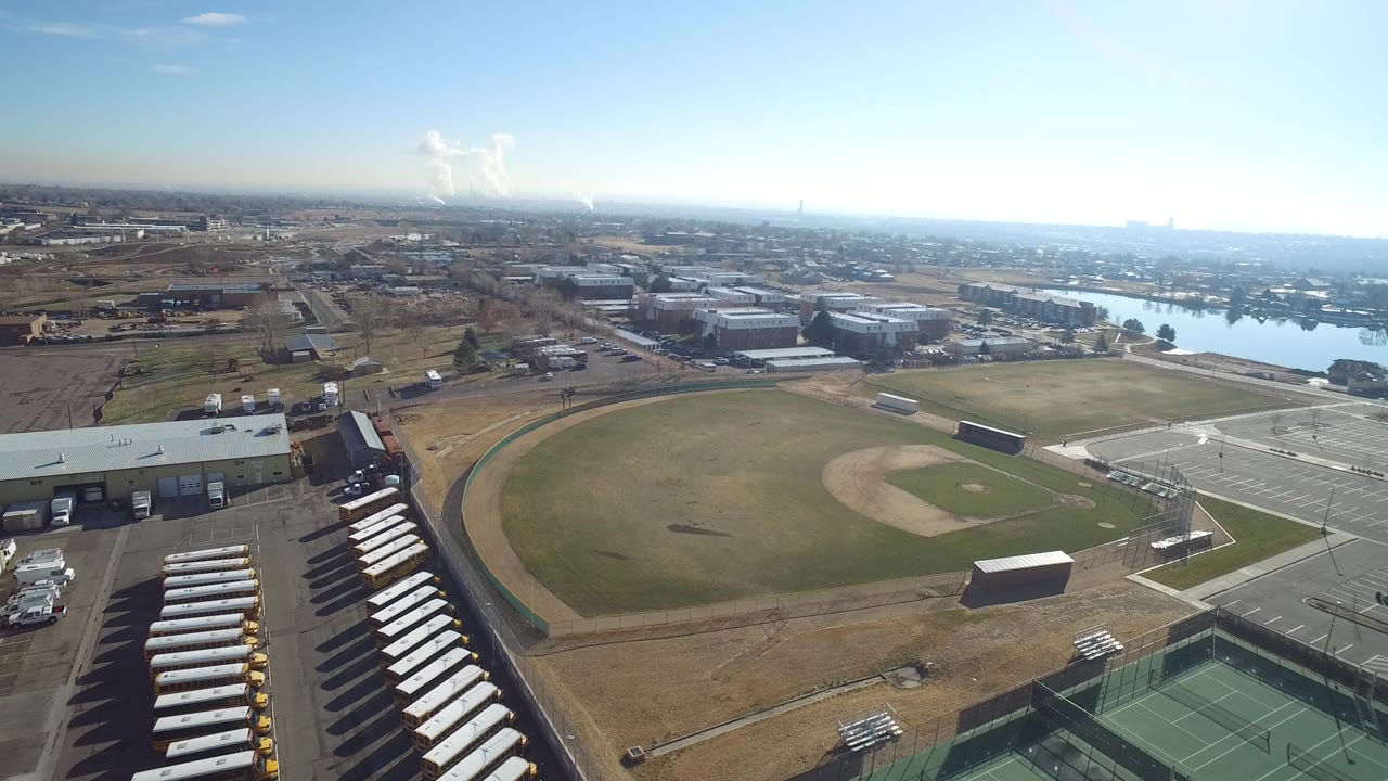 Aerial frame over Hidden Lake, Colorado showing more of the service buildings and parking lot beside the baseball field, with distant lake water and no visible people.