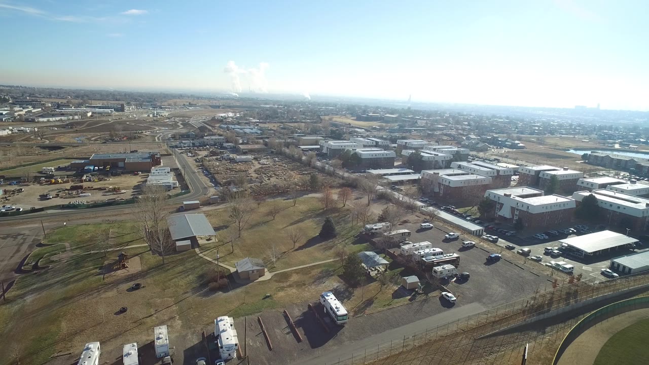 Drone view over Hidden Lake, Colorado showing open grass, parked vehicles, low-rise commercial buildings, roads, and a hazy skyline with no visible people.