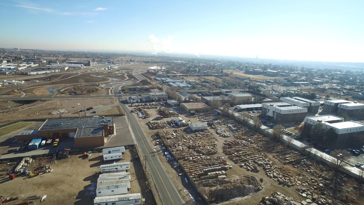 Wide aerial view over a Hidden Lake, Colorado car scrapyard with rows of broken vehicles, warehouse roofs, a straight road, distant ramps, and no visible people.