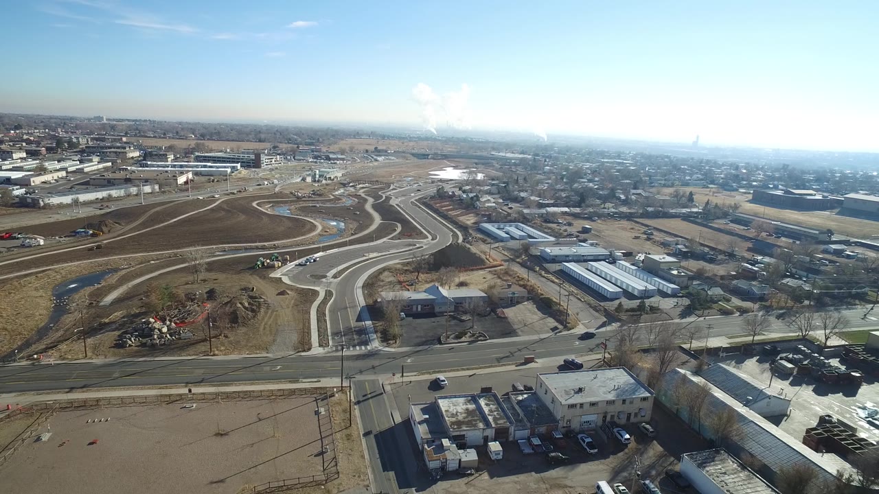 Wide aerial over a roadway junction with homes, low commercial buildings, and distant skyline.