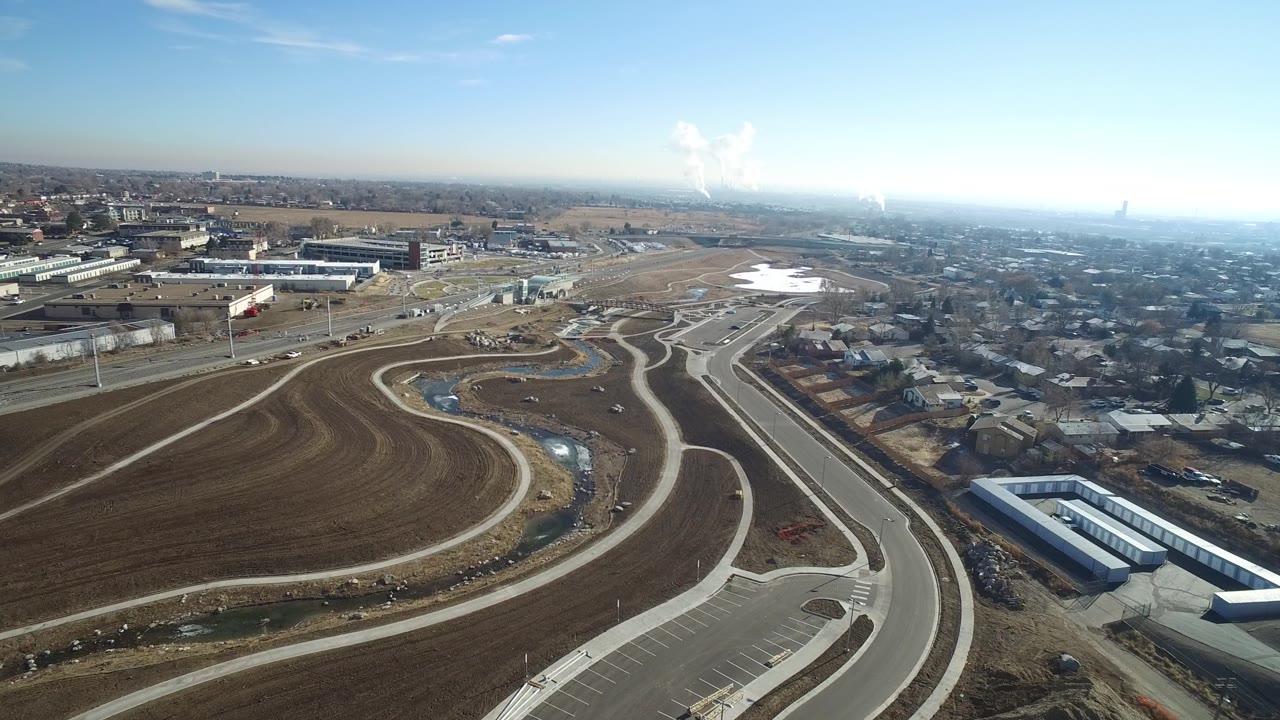 High aerial frame of graded land, winding streets, storage buildings, homes, and distant city haze.