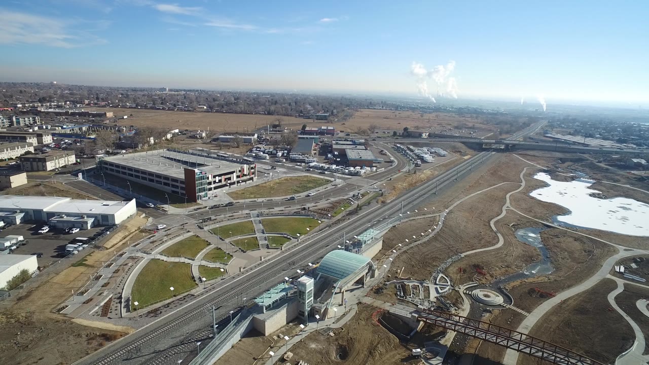 Second aerial frame over Hidden Lake, Colorado with the road and rail corridor running diagonally past low commercial buildings, paved paths, open land, snow patches, and no visible people.